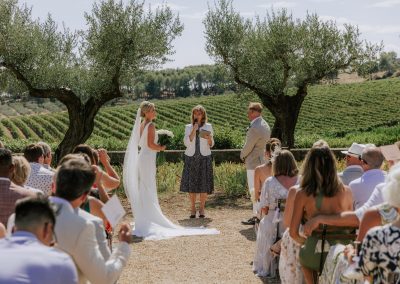 Bride in white, English speaking celebrant and groom stand with a backdrop of olive trees and vineyards in France