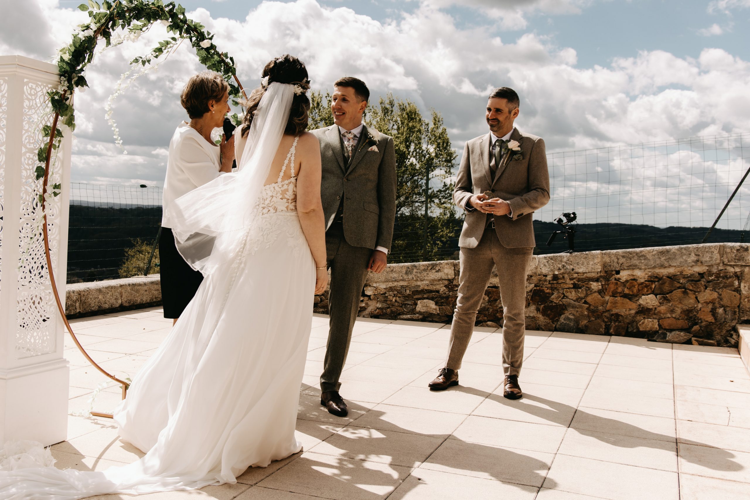 Bride in white dress and veil has her back to the camera, celebrant in white jacket and blue dress speaks to groom who wears a tweed suit